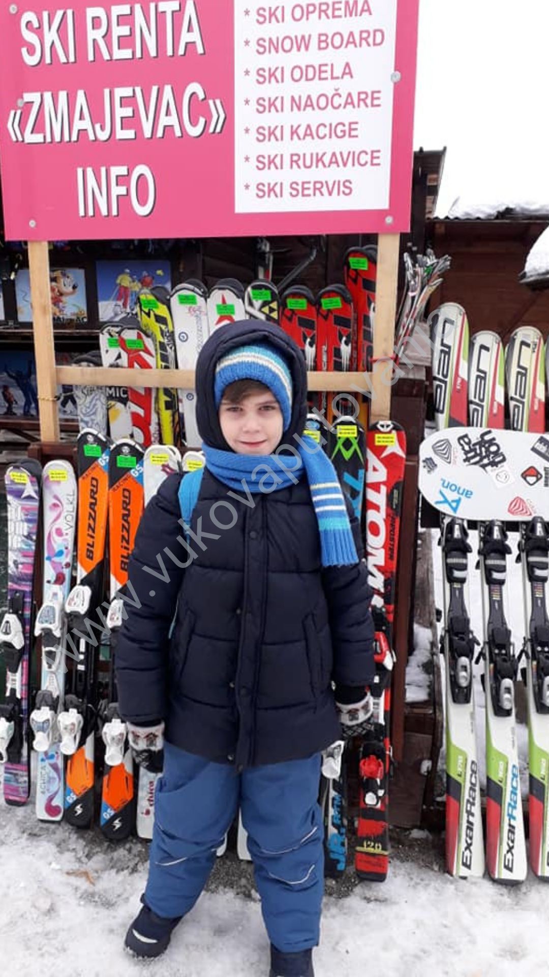 F.blog1 dečak u perjanoj teget jakni sa šalom i kapom stoji ispred ski opreme